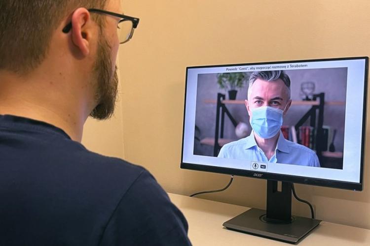 Zdjęcie mężczyzny przed ekranem monitora, na którym widoczna jest twarz innego mężczyzny w maseczce