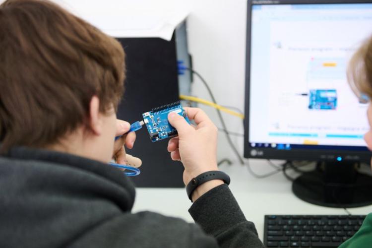 Zdjęcie ekranu komputera i siedzącej przed nią osoby trzymającej płytkę elektroniczną
