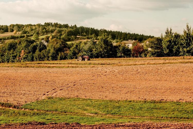 Na zdjęciu wiejski krajobraz: na pierwszym planie pole, w tle ciągnik, las i dom
