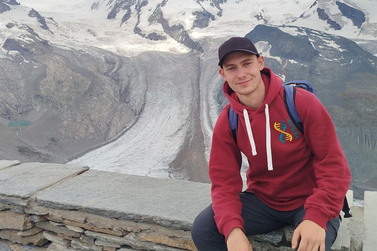 Na zdjęciu młody mężczyzna siedzi na murku, w tle widać góry - Alpy