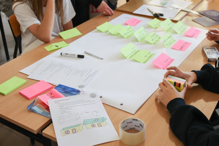 Na zdjęciu stół z rozłożonymi białymi kartkami i kolorowymi karteczkami oraz siedzące przy stole osoby