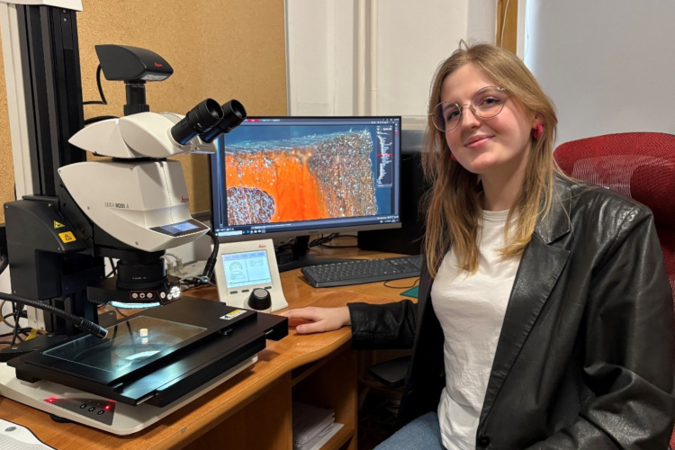 Kobieta siedzi przy biurku, uśmiecha się do zdjęcia, na biurku widać sprzęty naukowe.