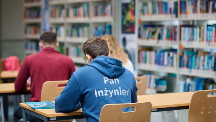 Na zdjęciu trzy osoby siedzące w bibliotece, widoczne tyłem 