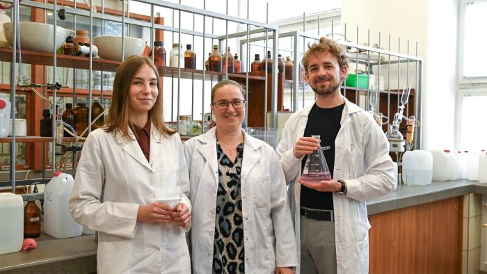 Na zdjęciu trzy osoby: dwie kobiety i mężczyzna, ubrani w fartuchy, stoją w laboratorium chemicznym