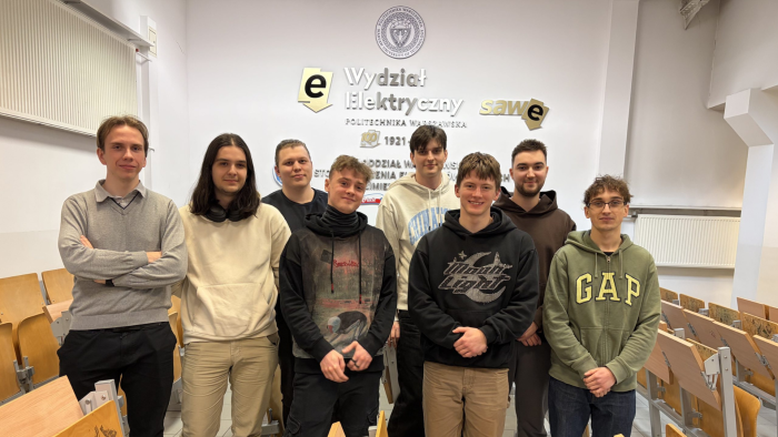 Zdjęcie przedstawia ośmiu studentów stojących na tle ściany z logotypem Wydziału Elektrycznego PW