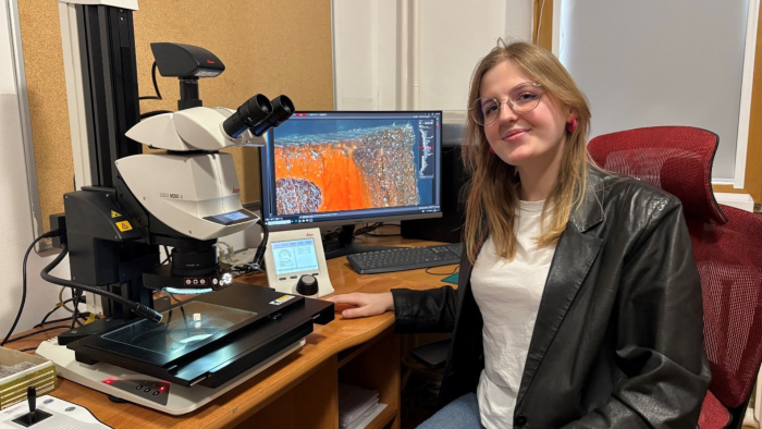 Kobieta siedzi przy biurku, uśmiecha się do zdjęcia, na biurku widać sprzęty naukowe.