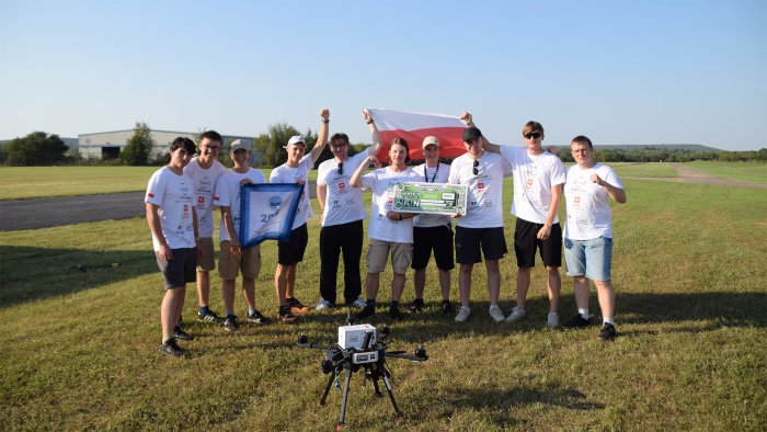 Na zdjęciu grupa dziesięciu studentów - stoi na otwartej przestrzeni, a przed nimi leży dron