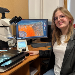 Kobieta siedzi przy biurku, uśmiecha się do zdjęcia, na biurku widać sprzęty naukowe.